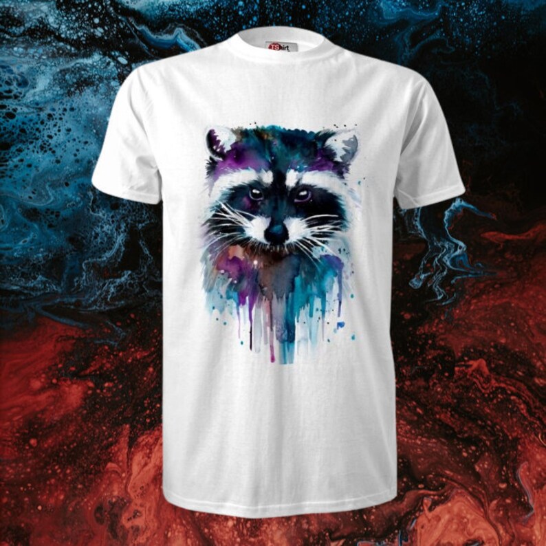 Op de afbeelding: Wit T-shirt met een aquarel-illustratie van een wasbeerkop. De vacht van de wasbeer is donkergrijs met witte markeringen. De achtergrond is een blauwe en paarse aquarel-wassing.
