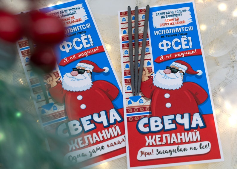 Puede incluir: Dos tarjetas rojas y azules con un Pap&aacute; Noel de dibujos animados que lleva gafas de sol. Las tarjetas tienen el texto "SVECHA ZHELANIY" y "Зажигай не только на танцполе! Зажигай свечу желаний! Исполнится! Не только лишь ФСЁ! Я не хадный!"  Las tarjetas est&aacute;n al lado de un paquete de bengalas plateadas.