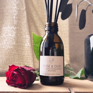 May include: A brown glass bottle with a black label that reads "Rose & Oud", "oud rose sandalwood", and "Reed Diffuser | 125ml". The bottle is filled with a clear liquid and has black sticks sticking out of the top. A red rose is in the foreground.