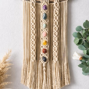 May include: Macrame wall hanging featuring a natural wooden dowel and beige cotton cord. The design includes a central column with seven colorful gemstones, and two decorative side columns. The bottom of the piece has a fringe.