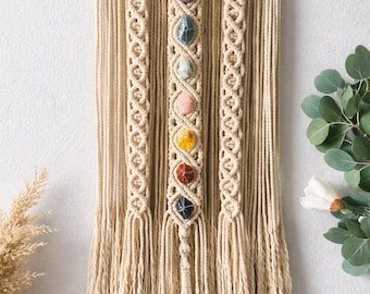 May include: Macrame wall hanging featuring a natural wooden dowel and beige cotton cord. The design includes a central column with seven colorful gemstones, and two decorative side columns. The bottom of the piece has a fringe.