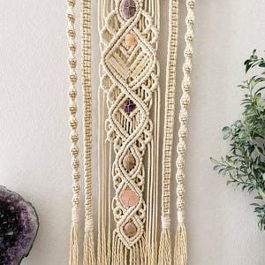 May include: Macrame wall hanging with a natural wood branch, featuring intricate woven patterns and embedded gemstones in shades of purple and pink. The off-white cotton fiber design includes knotted details and tassels, creating a bohemian aesthetic.