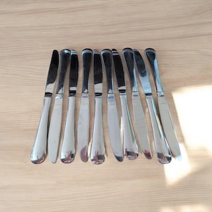 May include: A set of ten silver butter knives with rounded handles. The knives are arranged in a row on a wooden surface.