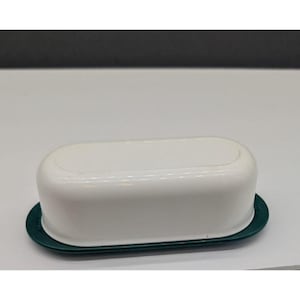 Puede incluir: Una mantequera rectangular blanca con la parte superior redondeada y una base verde oscuro. El plato está hecho de un material brillante y está diseñado para contener un tamaño estándar de mantequilla. La tapa encaja perfectamente en la base.