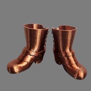 May include: A pair of miniature copper-colored boots. Each boot features a rounded toe, a strap across the ankle, and a mid-height heel. The boots are set against a solid gray background, showcasing their detailed design.