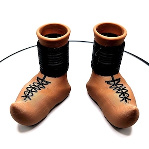 Puede incluir: Un par de botas de cerámica de terracota con cordones negros y una banda negra alrededor de la parte superior de cada bota.