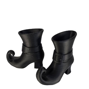 Puede incluir: Un par de botas de bruja negras con puntas puntiagudas y tacones pequeños. Cada bota tiene una correa con una hebilla alrededor del tobillo. Las botas son de color negro oscuro mate.