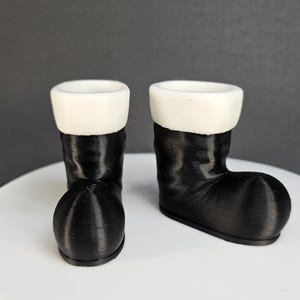 Könnte beinhalten: Zwei schwarze Nikolausstiefel mit weißem Fellbesatz. Die Stiefel sind aus einem strukturierten Material gefertigt und stehen auf einer weißen Oberfläche.