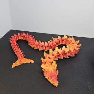 May include: A 3D-printed articulated dragon toy in shades of red and orange. The dragon is segmented, allowing for flexible posing. The head and tail are separate pieces. The dragon is on a black surface.