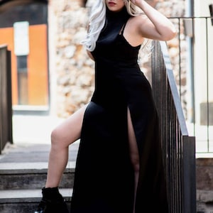 Puede incluir: Una mujer con cabello blanco y gafas de sol negras lleva un vestido negro maxi con una abertura en el muslo. Ella est&aacute; de pie en un conjunto de escaleras con una barandilla de metal negro.