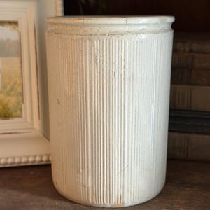 May include: A cylindrical off-white ceramic crock featuring a vertical ribbed texture along the exterior surface. The vessel has a smooth, slightly glossy finish with subtle signs of wear and ageing, including minor surface imperfections and cracks.