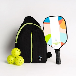 Könnte beinhalten: Ein schwarzer Rucksack mit neon-grünem Reißverschluss und einem kleinen Hunde-Logo. Vor dem Rucksack liegen drei gelbe Pickleball-Bälle. Rechts im Bild befindet sich ein bunter Pickleball-Schläger mit dem Text "swinton hero USA PICKLEBALL APPROVED".