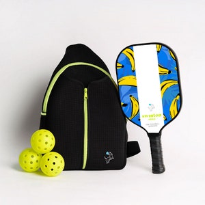 May include: A black and neon green pickleball bag with a zipper, a black pickleball paddle with a blue and yellow banana print, and three yellow pickleballs. The paddle has the text "Swinton Hero USA Pickleball Approved" on it.