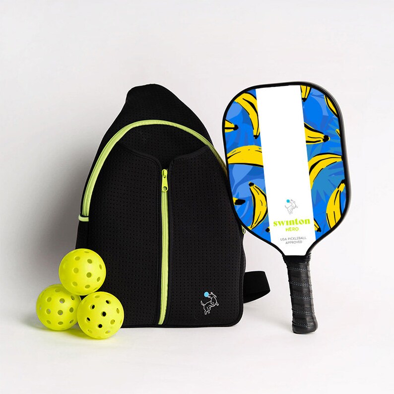 May include: A black and neon green pickleball bag with a zipper, a black pickleball paddle with a blue and yellow banana print, and three yellow pickleballs. The paddle has the text "Swinton Hero USA Pickleball Approved" on it.