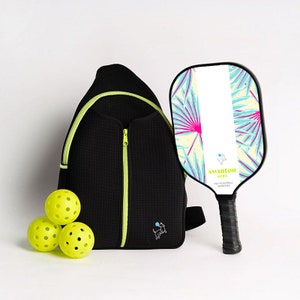 May include: A black and neon green pickleball backpack with a zippered pocket, a neon green dog logo, and three yellow pickleballs. A white and blue pickleball paddle with a tropical leaf design and the text "Swinton Hero USA Pickleball Approved" is also included.