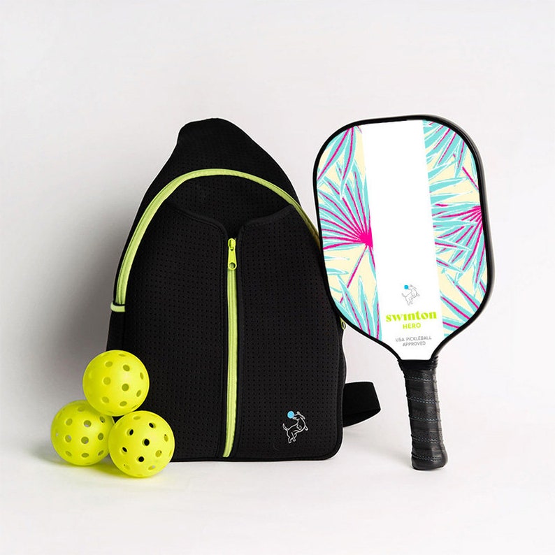 May include: A black and neon green pickleball backpack with a zippered pocket, a neon green dog logo, and three yellow pickleballs. A white and blue pickleball paddle with a tropical leaf design and the text "Swinton Hero USA Pickleball Approved" is also included.