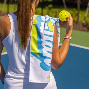 Puede incluir: Una toalla blanca con texto azul y amarillo que dice "Sweat Happens". Una persona est&aacute; sosteniendo una pelota de pickleball amarilla.