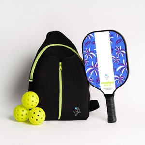 May include: A black pickleball backpack with a lime green zipper and a small dog logo. Three yellow pickleballs are in front of the bag. A blue and white pickleball paddle with a palm tree design and the text "swinton eclipse" is on the right side of the image.