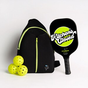May include: A black and green pickleball paddle with the text "Kitchen's Closed" in a white circle on a green background. The paddle is next to a black and neon green backpack with a small dog graphic. Three yellow pickleballs are in front of the backpack.
