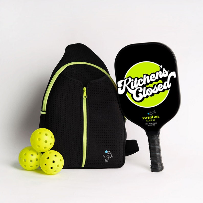 May include: A black and green pickleball paddle with the text "Kitchen's Closed" in a white circle on a green background. The paddle is next to a black and neon green backpack with a small dog graphic. Three yellow pickleballs are in front of the backpack.