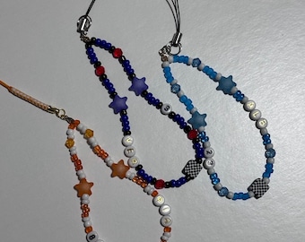 May include: Three beaded phone charms in blue, orange, and white. Each charm features star-shaped beads, letter beads, and a checkered square bead. The charms have black cords for attaching to a phone.