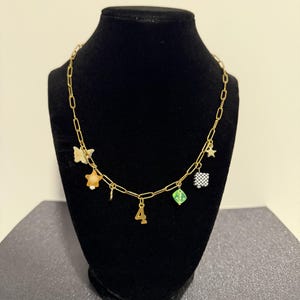 May include: A gold-toned chain necklace featuring multiple charms including a butterfly, a star, a number four, a green translucent cube, and a black and white checkered square. The necklace is displayed on a black stand to showcase the various dangling elements.