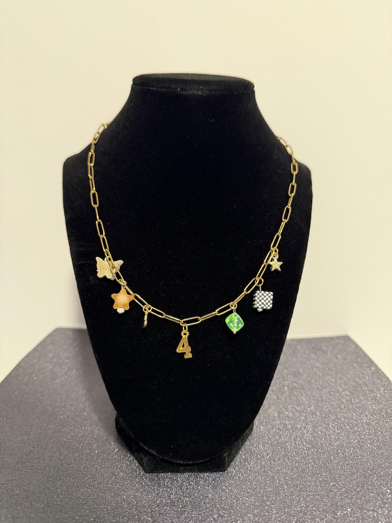 May include: A gold-toned chain necklace featuring multiple charms including a butterfly, a star, a number four, a green translucent cube, and a black and white checkered square. The necklace is displayed on a black stand to showcase the various dangling elements.