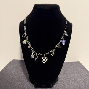 May include: A silver-toned chain necklace featuring various charms including a horseshoe, a white flower, the letters M and V, a black and white checkered heart, the number 3, a small heart outline, a purple star, and a small silver star.