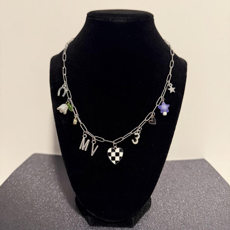 May include: A silver-toned chain necklace featuring various charms including a horseshoe, a white flower, the letters M and V, a black and white checkered heart, the number 3, a small heart outline, a purple star, and a small silver star.