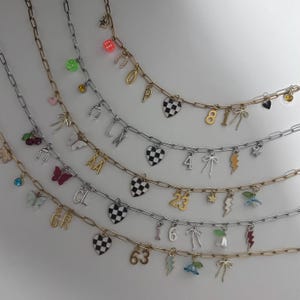 May include: Five link chain necklaces in gold and silver tones, each featuring an assortment of charms including checkered hearts, letters, numbers, bows, lightning bolts, flowers, butterflies, dice, horseshoes, and small colored crystals.