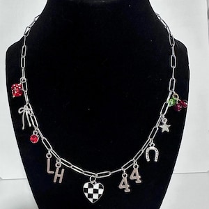 May include: A silver-toned chain necklace featuring various charms including a red die, a silver bow, a red crystal, the letters L and H, a black and white checkered heart, the number 44, a silver horseshoe, a silver star, and red cherries.