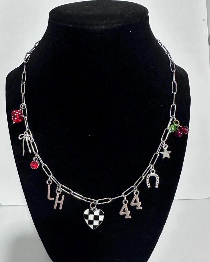 May include: A silver-toned chain necklace featuring various charms including a red die, a silver bow, a red crystal, the letters L and H, a black and white checkered heart, the number 44, a silver horseshoe, a silver star, and red cherries.