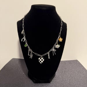 May include: A silver-toned chain necklace featuring various charms including a black and white checkered heart, the letters L and N, the number 1, a horseshoe, a green rhinestone, a small bow, a white flower, and a small orange star charm.