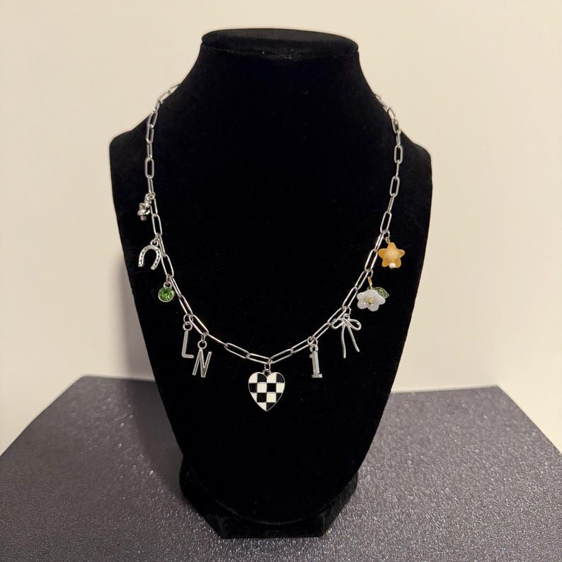 May include: A silver-toned chain necklace featuring various charms including a black and white checkered heart, the letters L and N, the number 1, a horseshoe, a green rhinestone, a small bow, a white flower, and a small orange star charm.