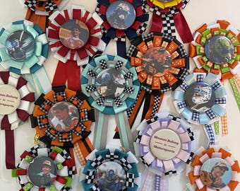 May include: A collection of circular fabric rosettes featuring various Formula 1 driver portraits and racing imagery. Each rosette is crafted with pleated ribbons in diverse colors and patterns, including checkered prints, with some displaying the text Women Belong in Motorsports.