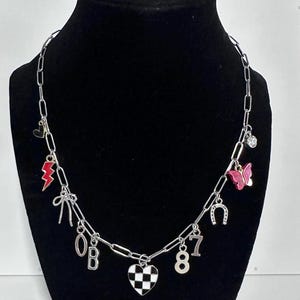 May include: A silver-toned chain necklace featuring various charms including a red lightning bolt, a pink butterfly, a horseshoe, a black and white checkered heart, a bow, a clear crystal, and the silver-colored characters 0, B, 8, and 7.
