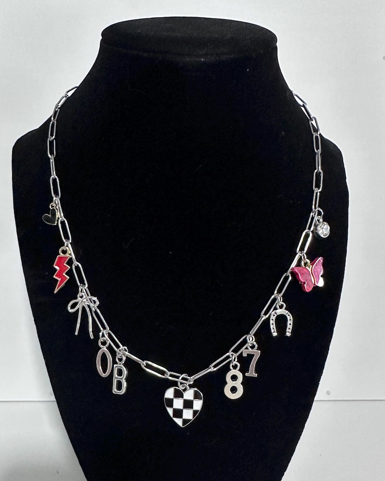 May include: A silver-toned chain necklace featuring various charms including a red lightning bolt, a pink butterfly, a horseshoe, a black and white checkered heart, a bow, a clear crystal, and the silver-colored characters 0, B, 8, and 7.