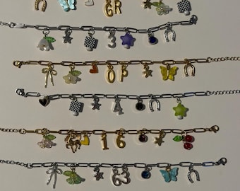 May include: A collection of seven charm bracelets featuring gold and silver-toned paperclip chains. Each bracelet is adorned with various colorful charms including butterflies, horseshoes, stars, hearts, flowers, cherries, and numbers or letters.