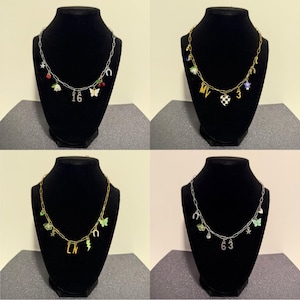 May include: Four silver and gold-toned chain necklaces featuring various charms including butterflies, horseshoes, stars, cherries, flowers, lightning bolts, hearts, and numbers 16, 63, and letters MV and LN, displayed on black jewelry busts.