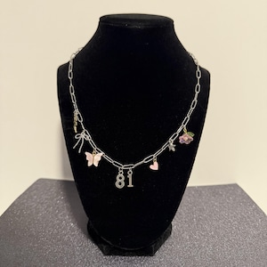 May include: A silver-toned chain necklace featuring multiple charms including a pink butterfly, a small pink heart, a silver bow, a small star, a flower with green leaves, and the numbers 8 and 1 hanging from the center of the link chain.