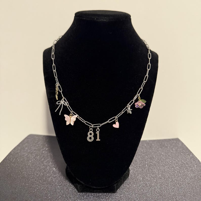 May include: A silver-toned chain necklace featuring multiple charms including a pink butterfly, a small pink heart, a silver bow, a small star, a flower with green leaves, and the numbers 8 and 1 hanging from the center of the link chain.