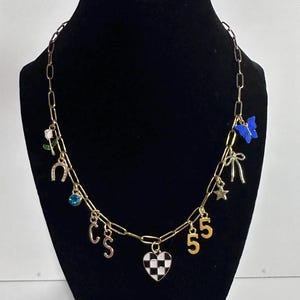 May include: A gold-toned chain necklace featuring various charms including a white rose, a horseshoe, a blue crystal, the letters C and S, a black and white checkered heart, the number 5, a star, a bow, and a blue butterfly charm.