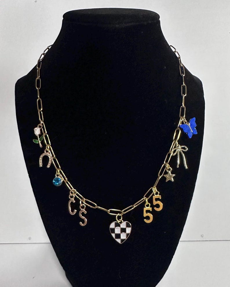 May include: A gold-toned chain necklace featuring various charms including a white rose, a horseshoe, a blue crystal, the letters C and S, a black and white checkered heart, the number 5, a star, a bow, and a blue butterfly charm.