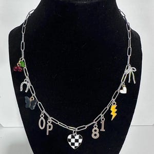 May include: A silver-toned chain necklace featuring various charms including red cherries, a horseshoe, a black butterfly, the letters O and P, a checkered heart, the numbers 8 and 1, a yellow lightning bolt, a small heart, and a silver bow.