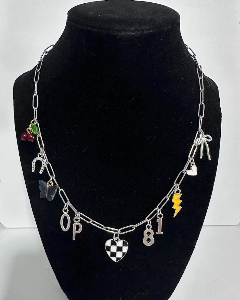 May include: A silver-toned chain necklace featuring various charms including red cherries, a horseshoe, a black butterfly, the letters O and P, a checkered heart, the numbers 8 and 1, a yellow lightning bolt, a small heart, and a silver bow.
