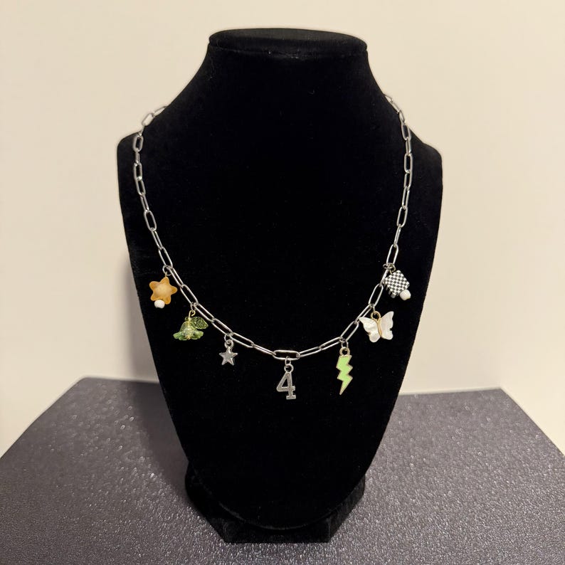May include: A silver-toned chain necklace featuring multiple charms including a yellow star, a green bell, a small silver star, a number four pendant, a green lightning bolt, a white butterfly, and a black and white checkered cube charm.