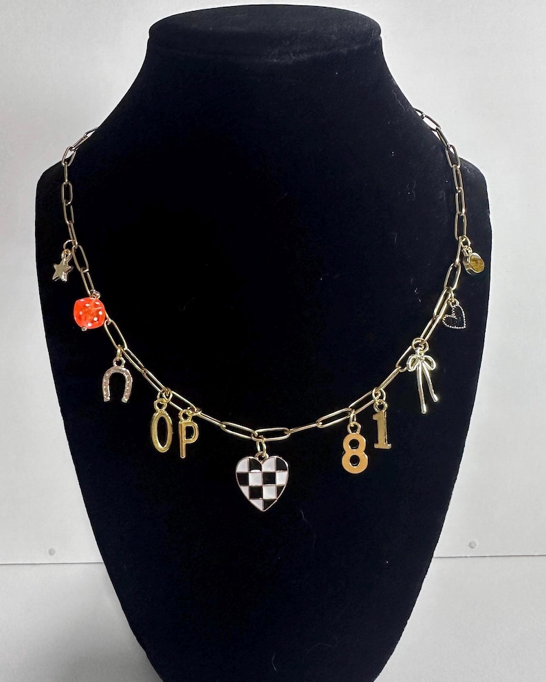 May include: A gold-toned chain necklace featuring various charms including a star, an orange die, a horseshoe, the letters O and P, a black and white checkered heart, the numbers 8 and 1, a bow, a small heart, and a yellow circular charm.