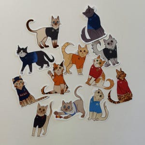 May include: A collection of twelve die-cut stickers featuring illustrated cats wearing various colored sports jerseys. The cats are depicted in different poses, some standing and some sitting, with several jerseys displaying small logos and numbers.