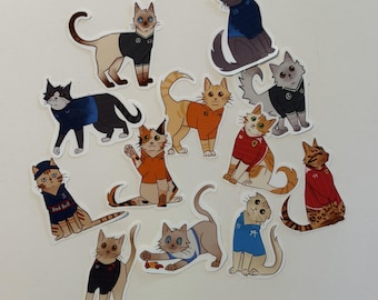May include: A collection of twelve die-cut stickers featuring illustrated cats wearing various colored sports jerseys. The cats are depicted in different poses, some standing and some sitting, with several jerseys displaying small logos and numbers.