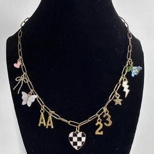 May include: A gold-toned chain necklace featuring various charms including a pink heart, a bow, a white butterfly, the letters AA, a black and white checkered heart, the numbers 23, a star, a lightning bolt, and a blue flower with a green leaf.